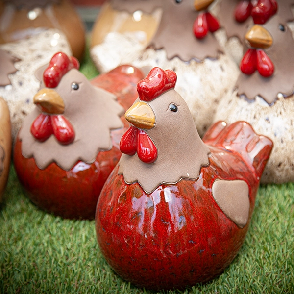 Sculptures de poules en céramique avec des plumes rouges et des crêtes jaunes. Les sculptures sont disposées sur une surface herbeuse.