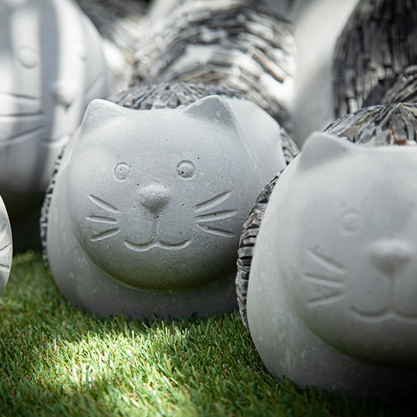 Sculptures de chats en céramique avec des visages souriants, disposées sur une surface herbeuse. Les sculptures sont de couleur gris clair et ont des détails texturés.
