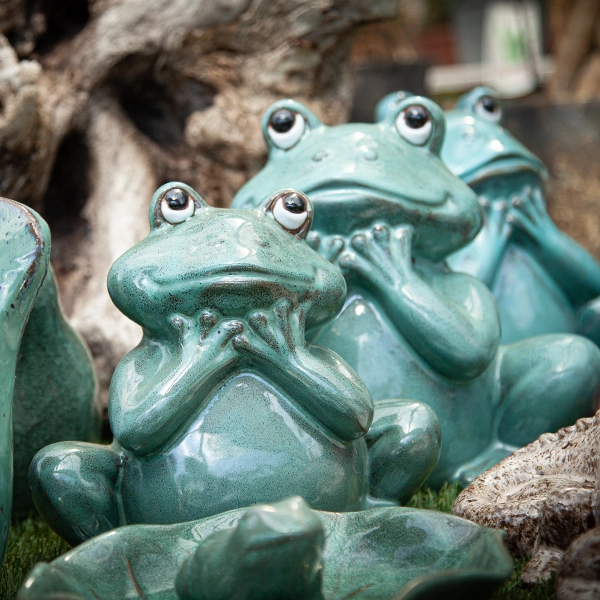 Sculptures de grenouilles en céramique avec des yeux mobiles, disposées sur des feuilles de nénuphar. Les sculptures sont de couleur verte et sont placées dans un environnement de jardin ou de serre.