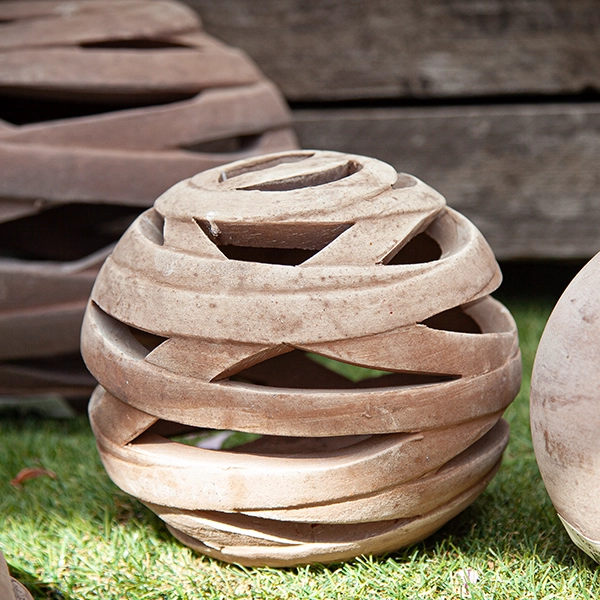 Sculpture sphérique en céramique avec des motifs en spirale, disposée sur une surface herbeuse. La sculpture est de couleur beige et a une texture rugueuse.