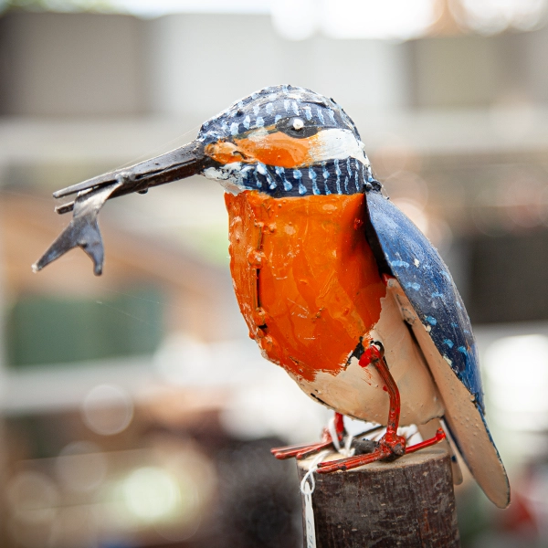 Sculpture d'un oiseau en métal avec des détails colorés, incluant une poitrine orange et des ailes bleues. L'oiseau est perché sur un piquet en bois, avec un poisson en métal dans son bec.