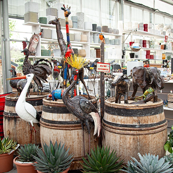 Une collection de sculptures d'animaux en métal, incluant des oiseaux, une girafe et un éléphant, disposées sur des barils en bois. Les sculptures sont entourées de plantes en pot, dans un environnement de serre ou de jardin.