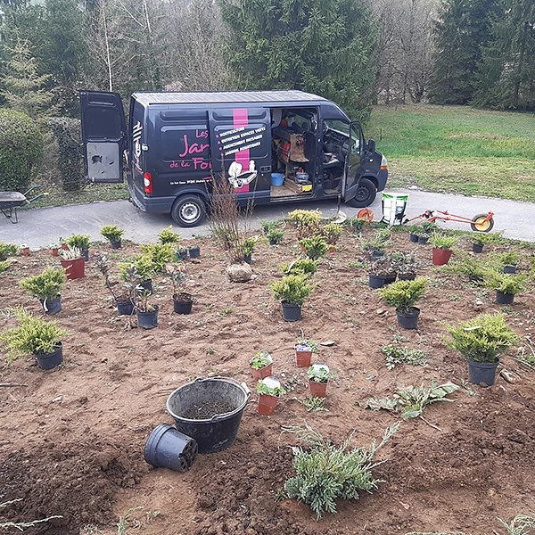 Le van des Jardins de la Fontaine est garé à côté d'un petit jardin avec des plantes en pot disposées en rangées. Le van a une porte ouverte et des outils de jardinage sont visibles à l'intérieur. Le jardin est entouré d'une zone herbeuse et d'arbres en arrière-plan.