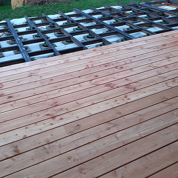 Une terrasse en bois en cours de construction. Au premier plan, des planches de bois sont posées et alignées. En arrière-plan, une structure de support en métal avec des blocs de béton est visible, indiquant la phase de construction de la terrasse.